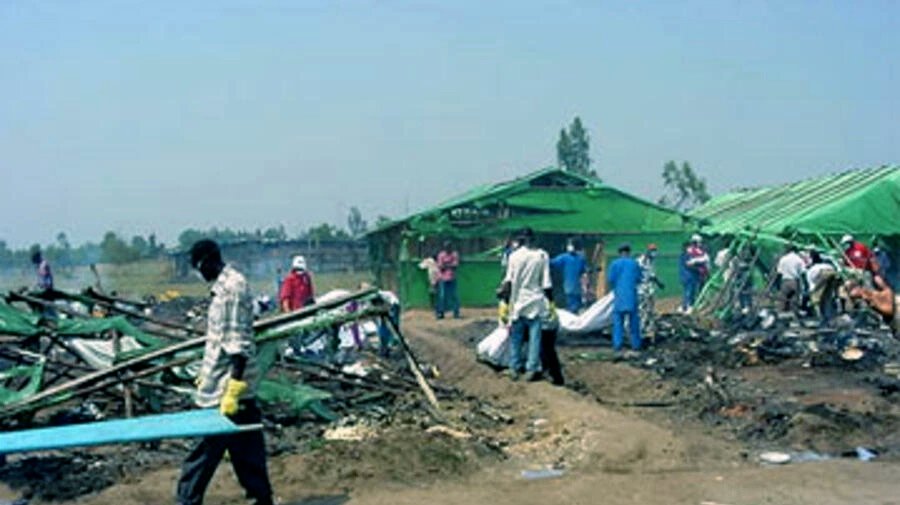 Destroyed green-tarped shelters and recovery workers at Gatumba after the massacre